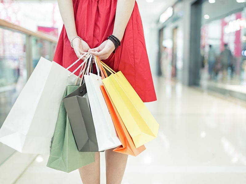 Mulher vestindo um vestido vermelho, segurando várias sacolas de compras de diferentes cores em um centro comercial iluminado e moderno.