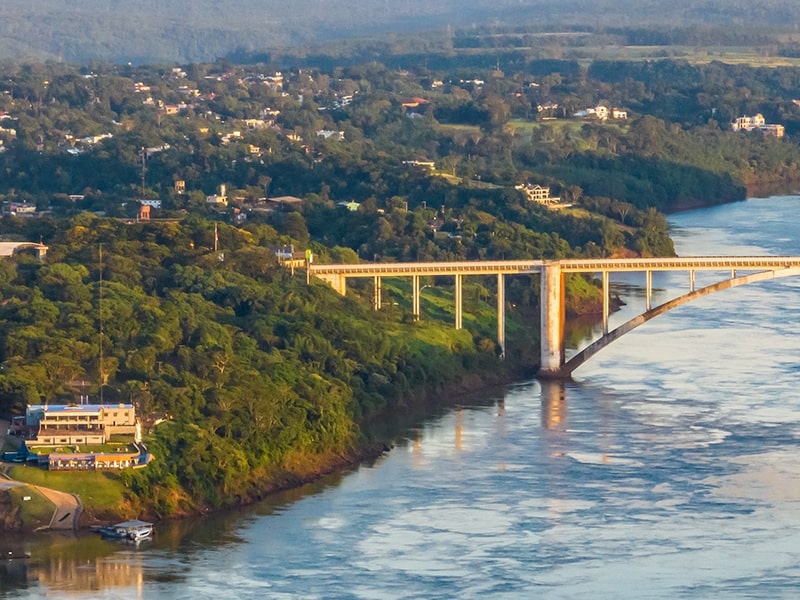 Vista aérea da Ponte da Amizade sobre o rio Paraná, conectando Foz do Iguaçu no Brasil a Ciudad del Este no Paraguai, destacando a vegetação exuberante e a paisagem urbana. Ideal para quem planeja viajar para o Paraguai a partir de Foz do Iguaçu.