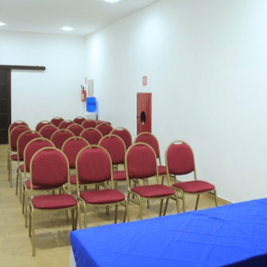 Sala de conferências com cadeiras vermelhas dispostas em fileiras, mesa coberta com toalha azul na frente.