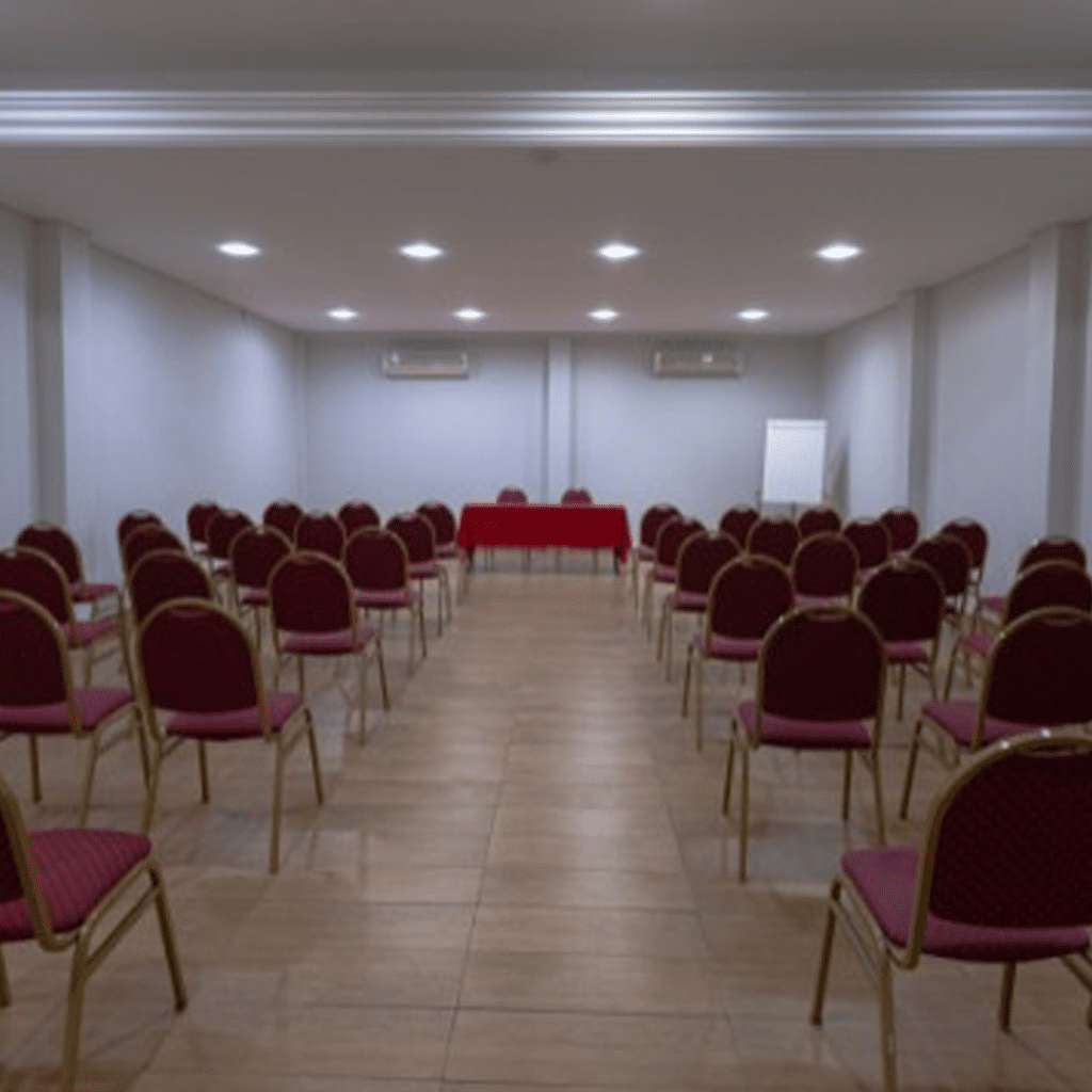 Espaço de eventos com cadeiras organizadas em fileiras simétricas, mesa coberta com toalha vermelha.