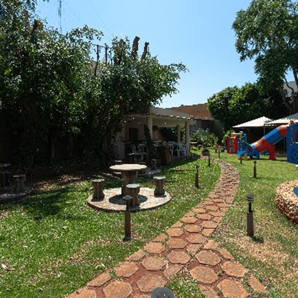 Jardim ao ar livre com mesas de concreto, caminho de pedras e área de playground.